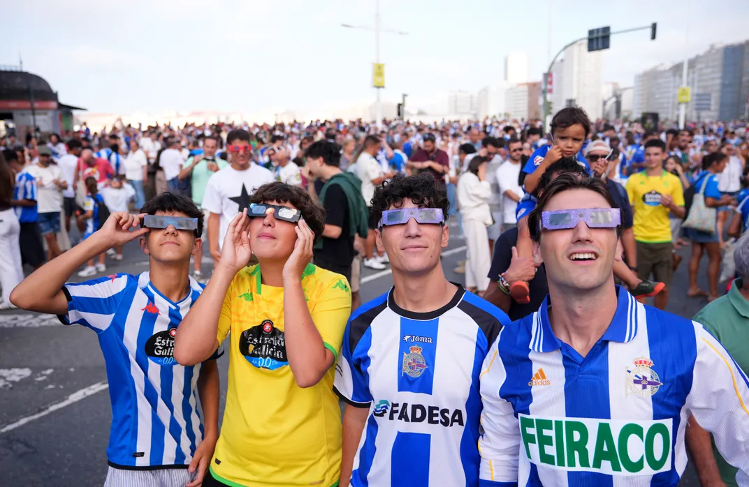 four young men in soccer jerseys look through eclipse glasses