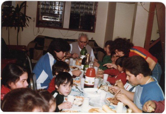 Family gathers around a table full of soda and dishes. An old man sits at the head of the table.