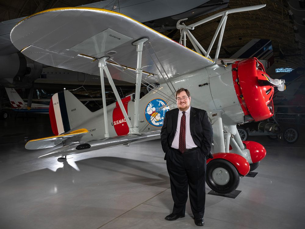 A white man in his 30s stands in front of a restored biplane displayed on a polished concrete floor inside a museum. He is wearing glasses and a dark suit with a white shirt and burgundy tie.