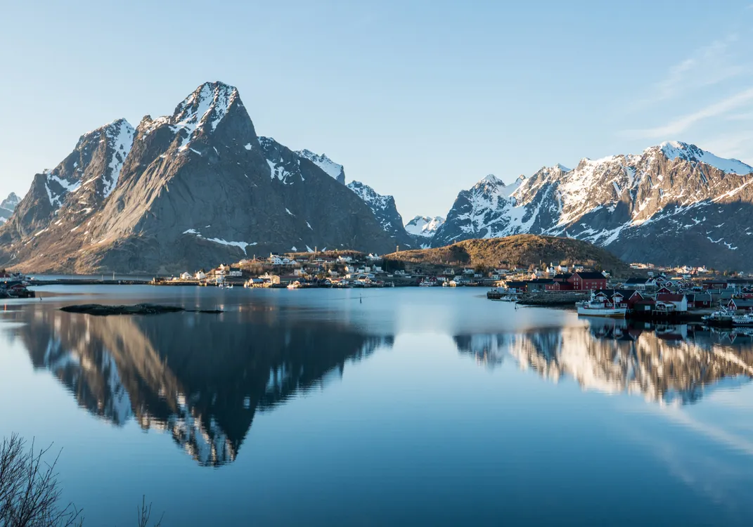 A calm lake reflects snow-capped mountains and a small village at their base under a clear, sunlit sky.