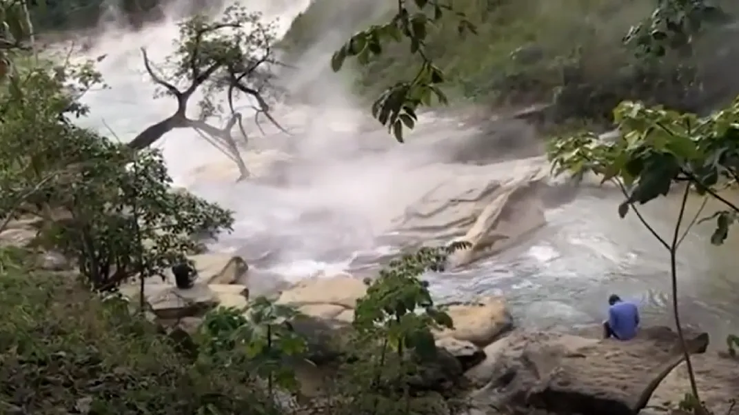 A man in a blue shirt sits on a rock near a section of the boiling river.