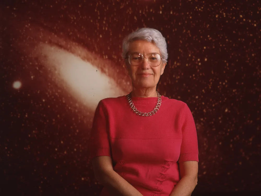 Vera Rubin stands against a backdrop of a star-filled galaxy with her hands loosely clasped in front of her.