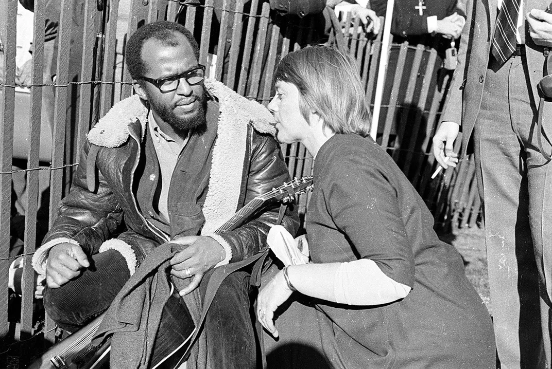 Woman talks with man in a leather jacket holding an acoustic guitar, both sitting on the ground alongside a picket fence.