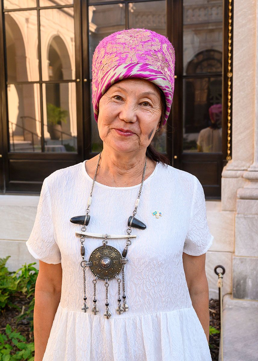 A woman with a cylindrical pink hat, white dress, and ornate metal necklace, smiles outdoors.