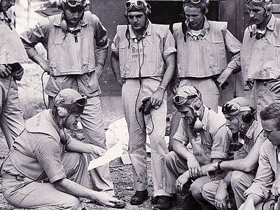 At the Black Sheep Squadron's base on the South Pacific island of Espiritu Santo, Boyington (holding paper) briefs his pilots on an upcoming mission.