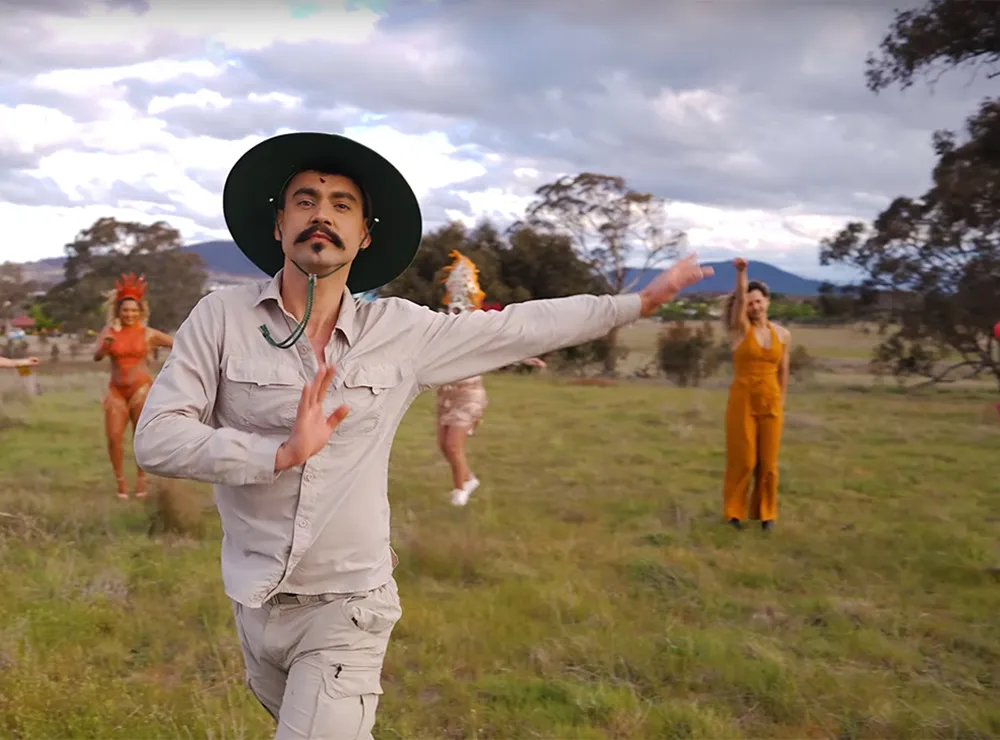Weliton Menário Costa dances in the foreground of a grassy plain, while background dancers dressed in orange dance behind him.