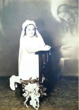 First communion photograph of a girl kneeling down with her hands together in prayer pose. In the background an image of a man with a halo around his head.Clear