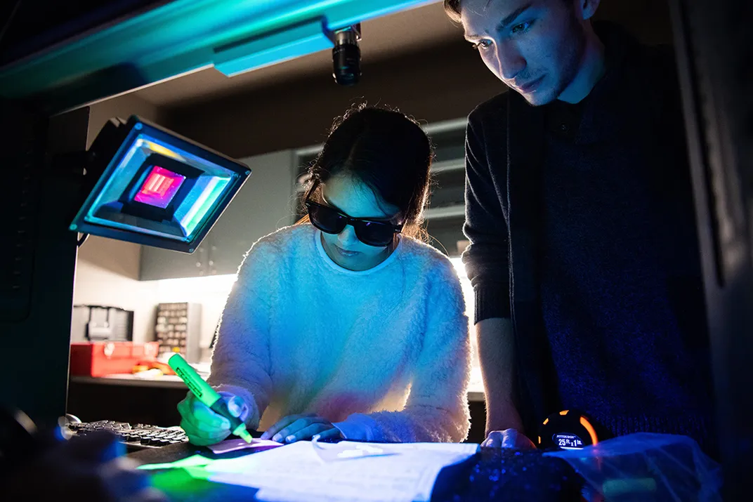 Two people stand overlooking a table; center, the person wears glasses and their face is illuminated blue as they highlight something on a piece of paper; they appear to be in a laboratory or classroom setting