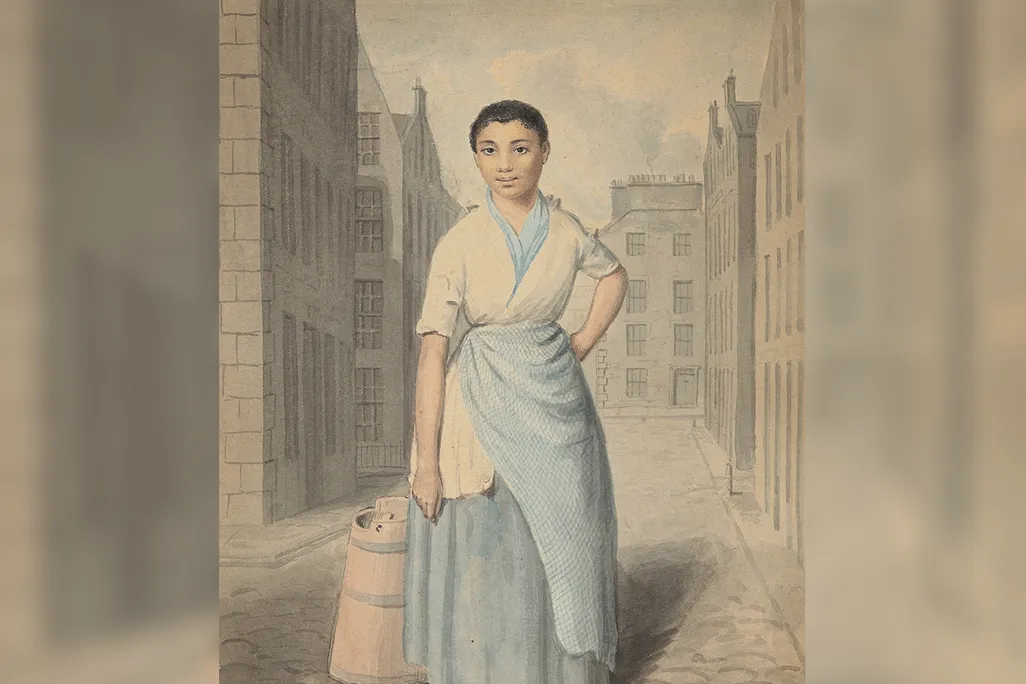 A solo full-length portrait of a Black woman in a white-and-blue wrapped dress, with cropped hair, standing with one hand on hip and barefoot on a cobbled street