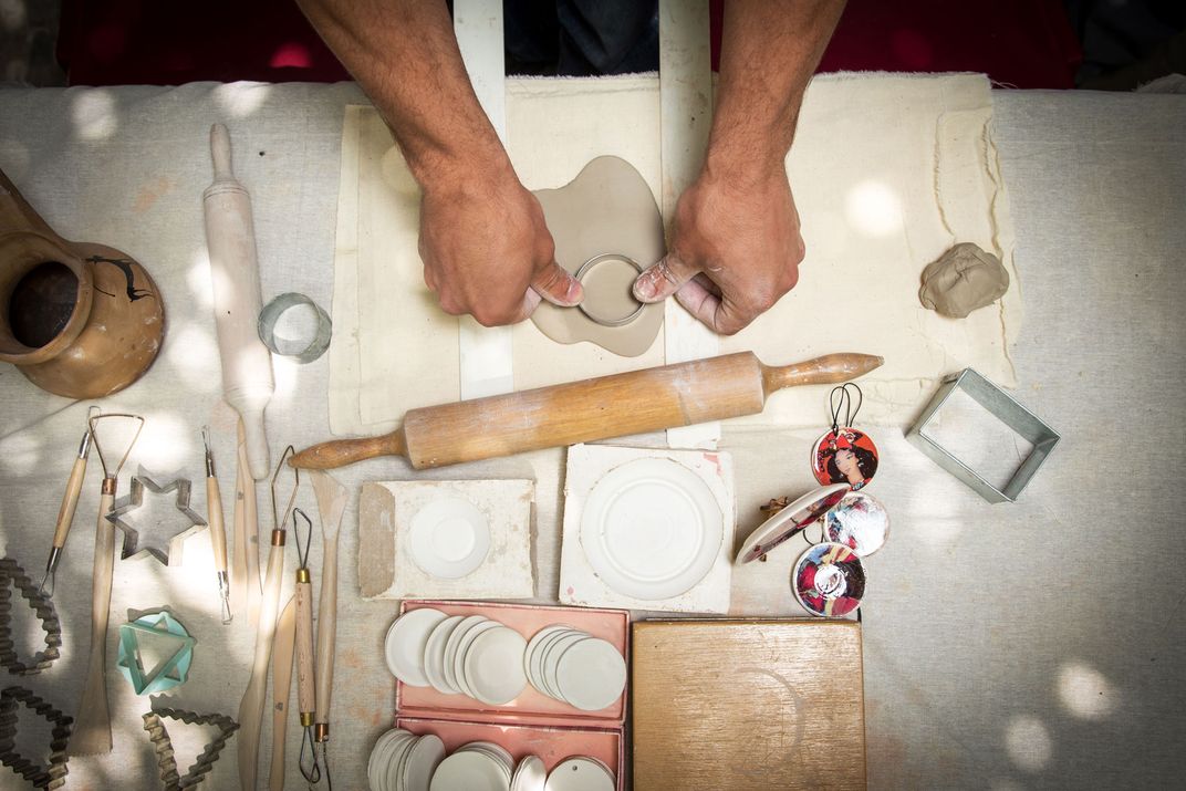 Spread out on a table are a variety of pottery tools and shape cutters. In the middle of the table is a rolled out piece of clay about to be cut with a circular shape.