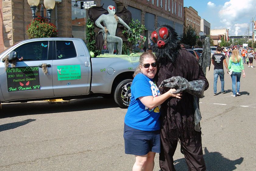 At an outdoor street festival, a woman embraces a person dressed us in a dark fuzzy Mothman costume, with a dark mask with red eyes and mouth.