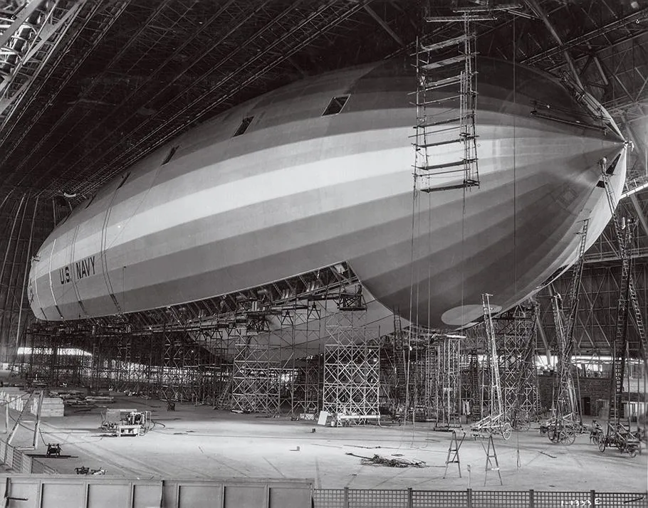 A black-and-white photo shows a silver-colored airship inside a hangar, with a few struts and girders visible on the front half.
