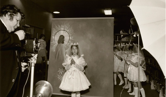 Black and white photograph of a girls posing for a first communion picture. A dropdown background image of a man with a halo around his head. A photographer stands with a camera in front of the girl.