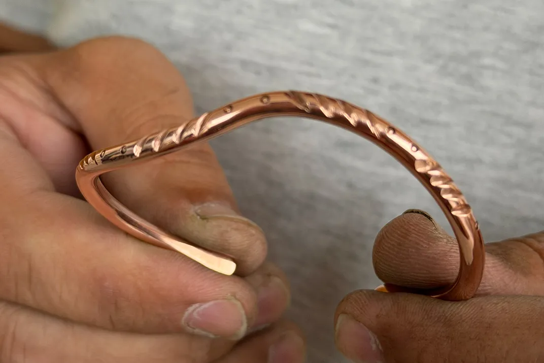 Close-up on a pair of hands holding up a bracelet made of a thin, curving piece of copper, with diagonal lines and dots etched into it.