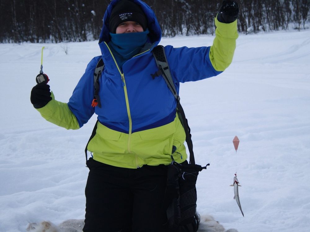 Person wearing winter gear kneeling in the snow holding up a fishing line with a small fish hooked on it