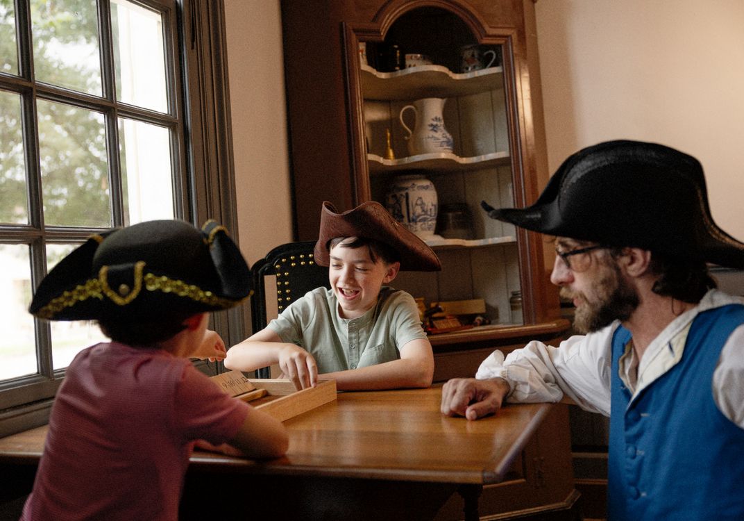 Visitors seated around a wooden table inside Shields Tavern in Colonial Williamsburg participate in a colonial-era activity with an interpreter in period clothing. Historic furnishings and decorative ceramics fill the tavern interior.