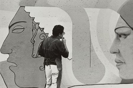 Grayscale image of Emanuel Martínez in a dark colored shirt and apron and light coloroed pants pondering a wall mural with two large faces.