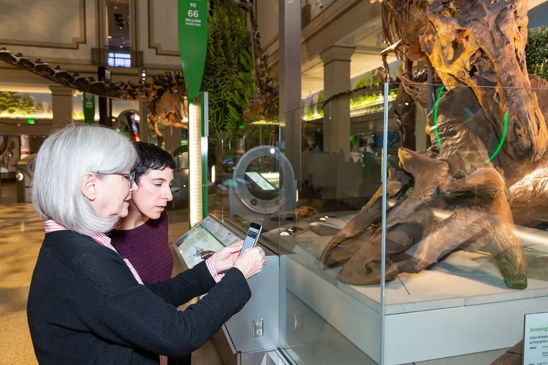 Two women looking at the Deep Time Audio Description app on a smartphone while standing in front of a display featuring a T. rex decapitating a triceratops in the Smithsonian's new fossil hall. 