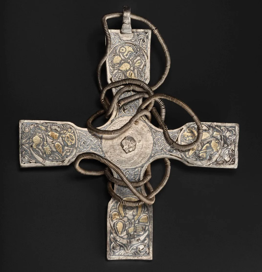 A silver cross, with a wire that coils around its surface and four equally long arms engraved with delicate depictions of a Human, Cow, Lion and Eagle