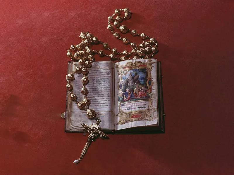 On a red background, an open illuminated Bible with colorful illustrations depicting Jesus kneeling and praying, encircled by a golden rosary with large round beads and a cross decorated with pearls and a golden figure of Jesus crucified on its end