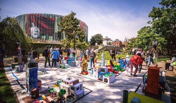 A large outdoor space is filled with human-sized colorful cardboard structures. In the background, the Hirshhorn Museum and Smithsonian Arts and Industries building are visible.