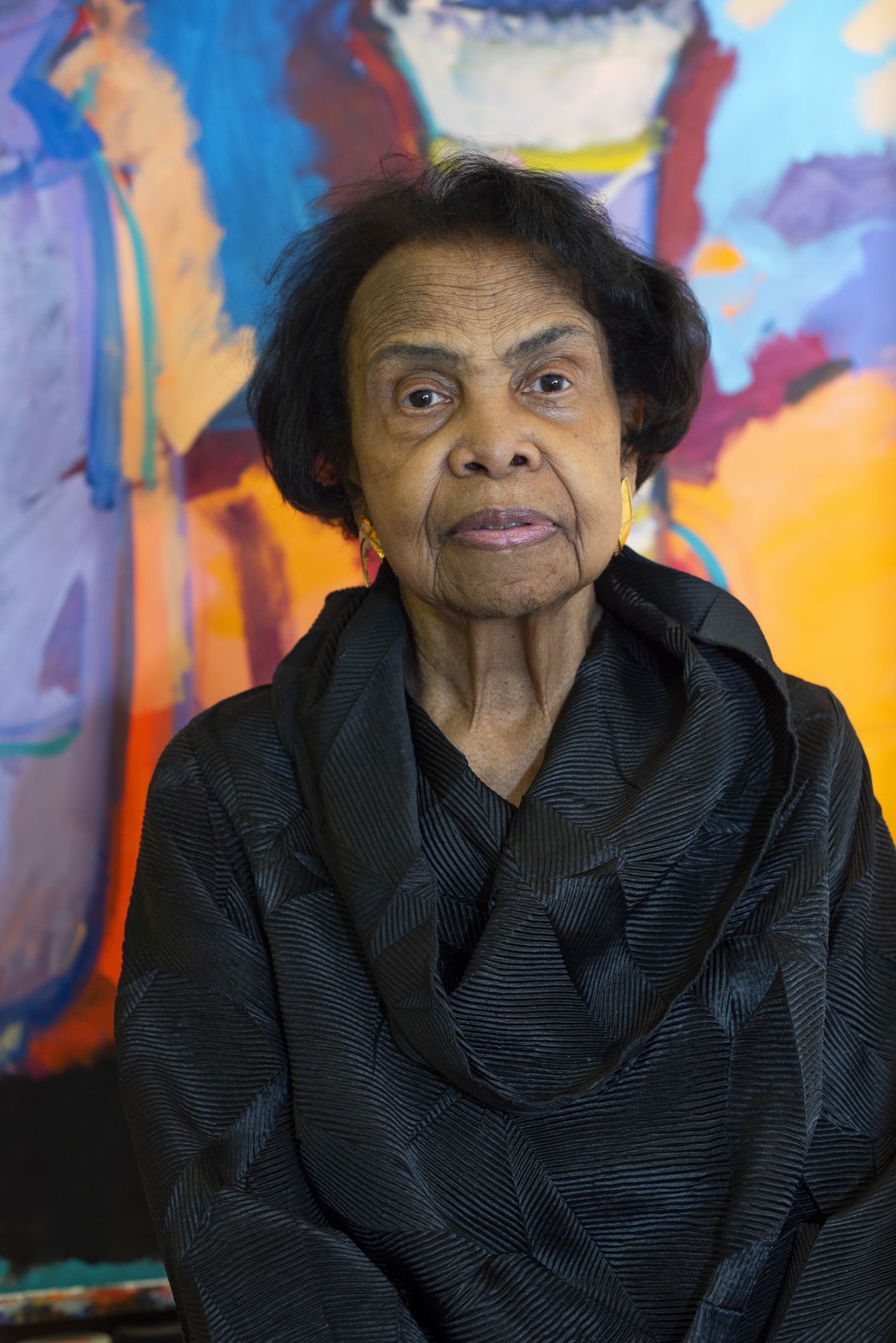 A picture of Woodson, a Black woman in her 80s, standing in front of a colorful canvas and wearing a black blouse