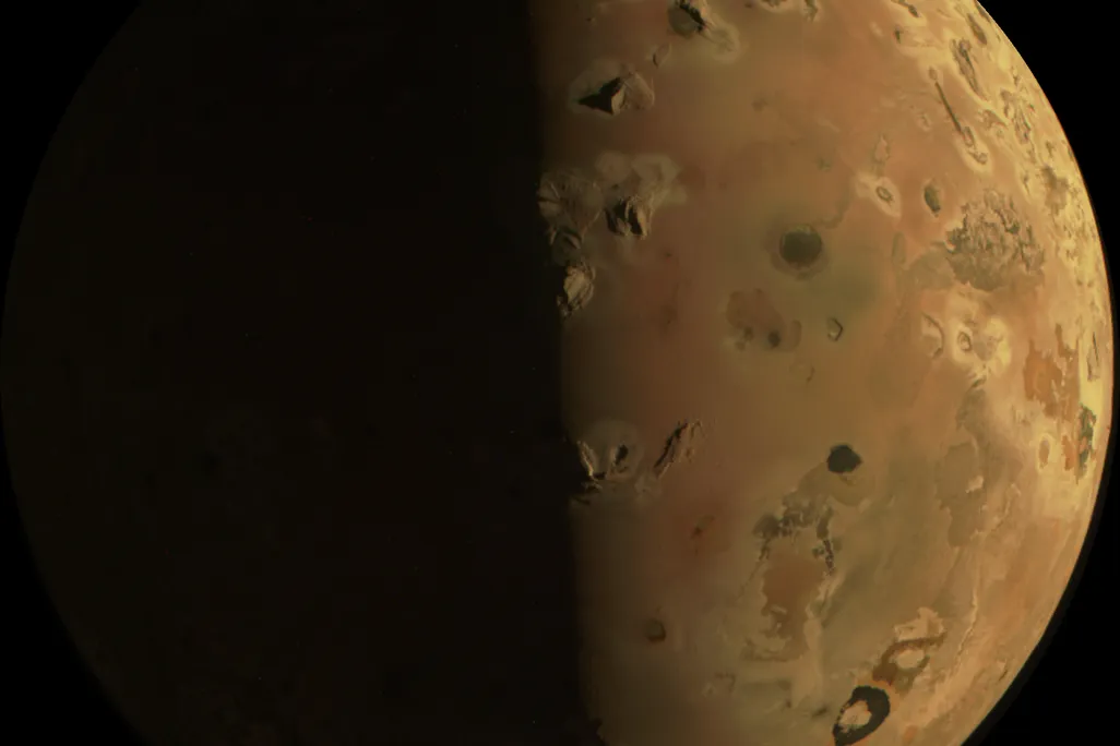 An image of Io, Jupiter's third-largest moon, appearing rust-colored with many visible black pockmarks, which are mountain ranges and surface volcanoes as seen from afar.