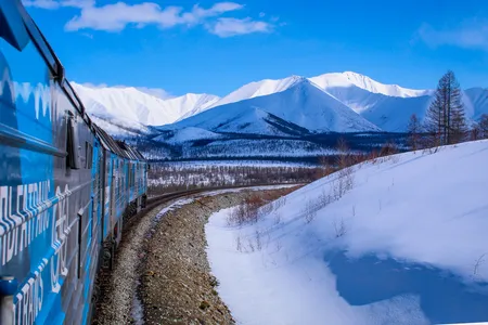 A train whizzes through snowy hills with frosty peaks from the Stanovoy Ridge mountains in the distance.