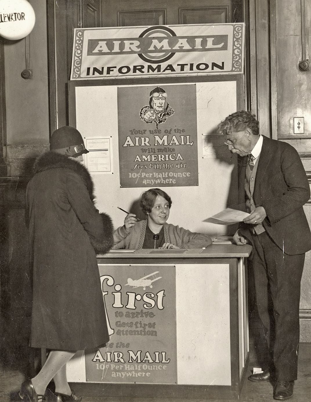 A woman in a hat stands on the left of an air mail booth where a woman sits behind the desk holds a pencil and looks at a man in a suit holding a piece of paper.