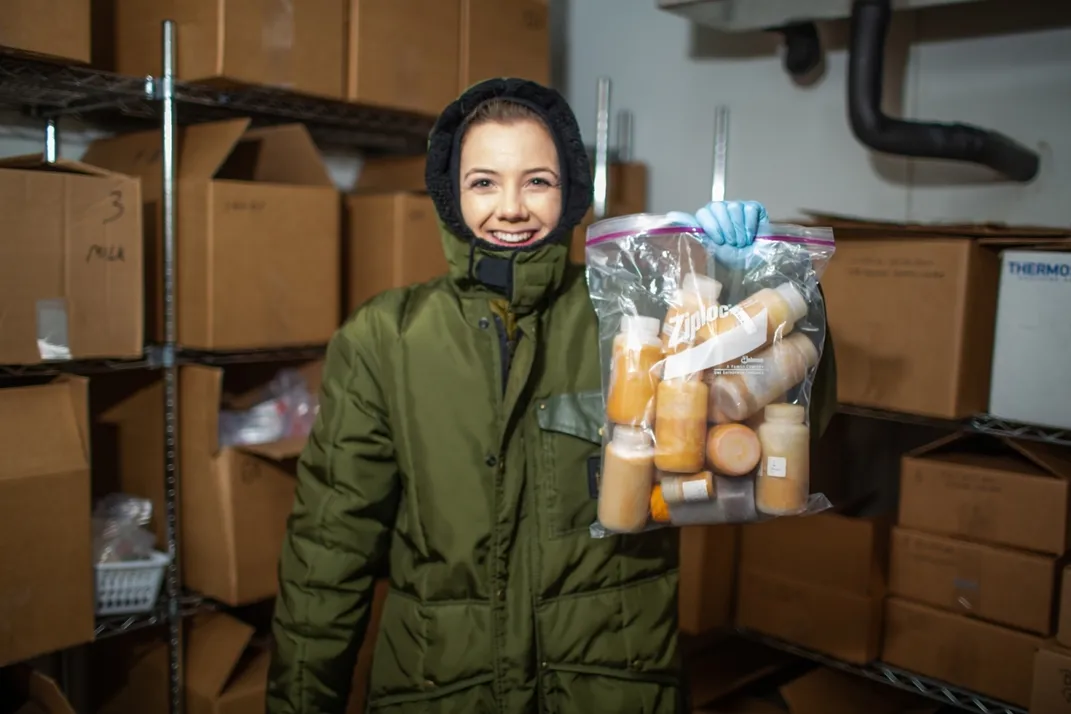 Nutrition lab research assistant Jenna Pastel stands inside the freezer that contains the animal milk repository. She's wearing full-body, hooded snowsuit and holding a bag of bottles of orange-colored animal milks.