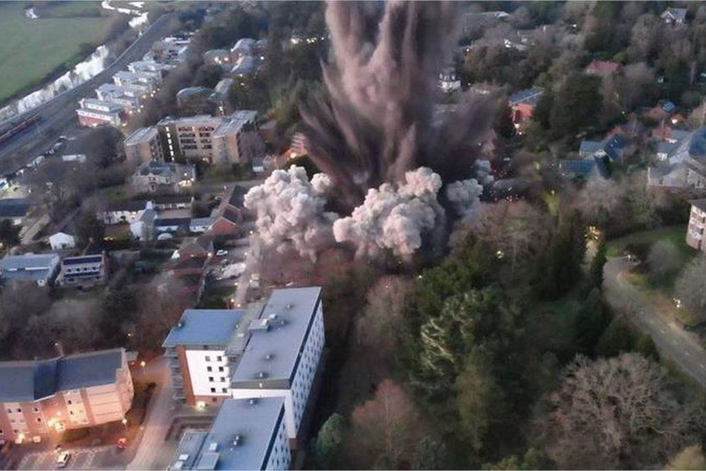 An aerial view of a huge explosion taking place in the middle of a neighborhood, with dust and smoke clouds emerging from buildings and surrounded by green trees