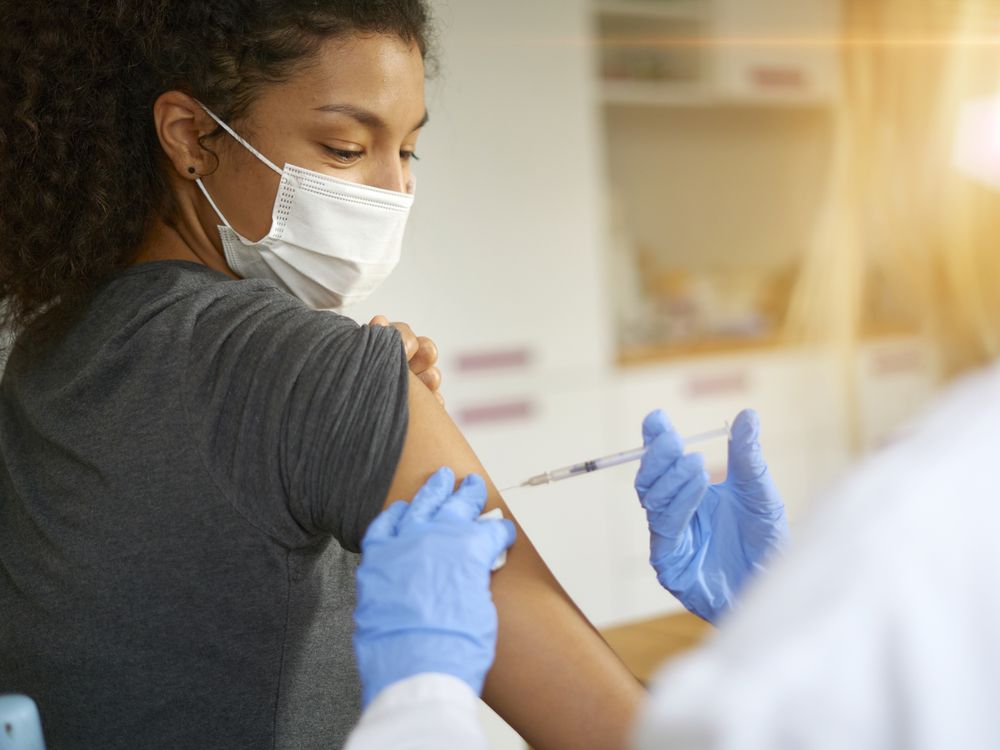 a woman in a mask gets a vaccine in her arm from a person in a lab coat and gloves