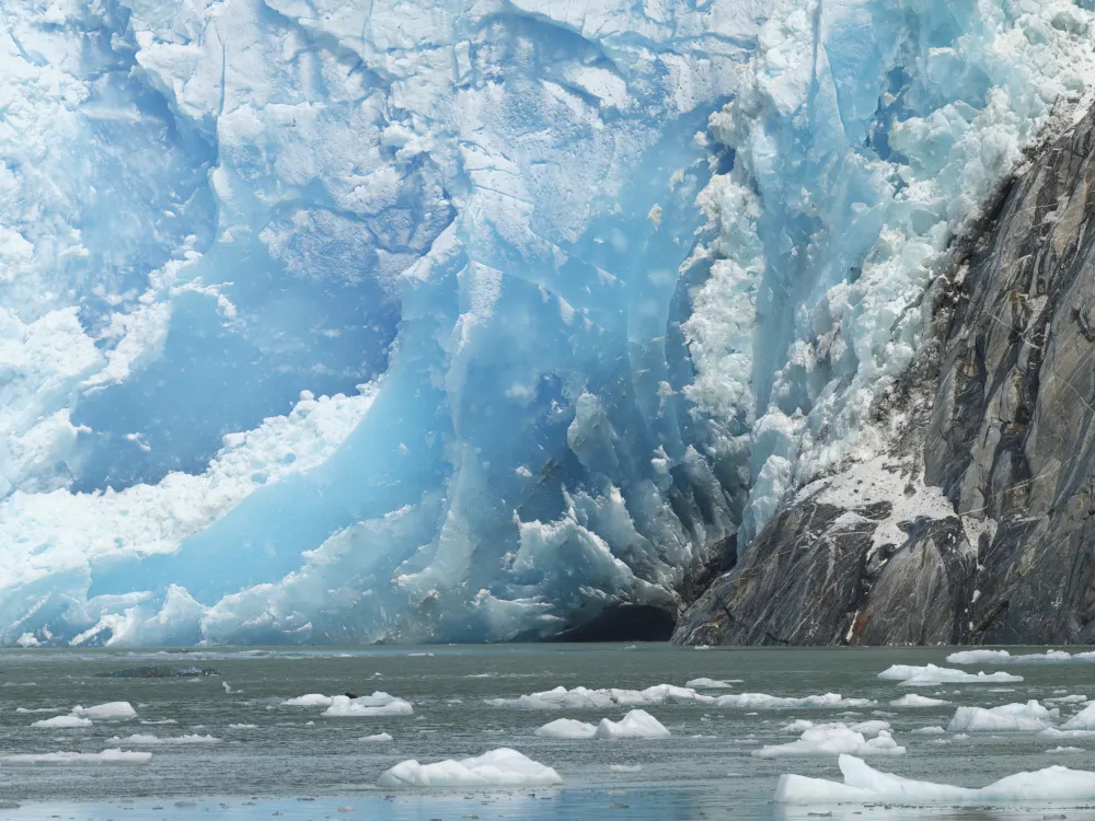 The base of South Sawyer Glacier forms a wall of blue glacial ice and grey rock, foregrounded by the waters of Tracy Arm, dotted by icebergs.