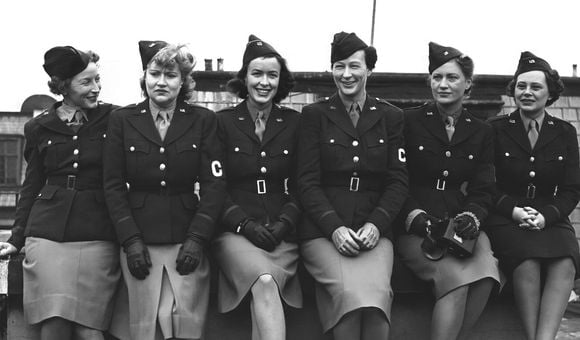 Six women sitting closely together posing for the camera. They are wearing uniforms with light skirts and dark jackets and an armband with the letter C on it.