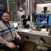 A man in a grey short sits in a lab setting, in front of a microscope and a computer screen.