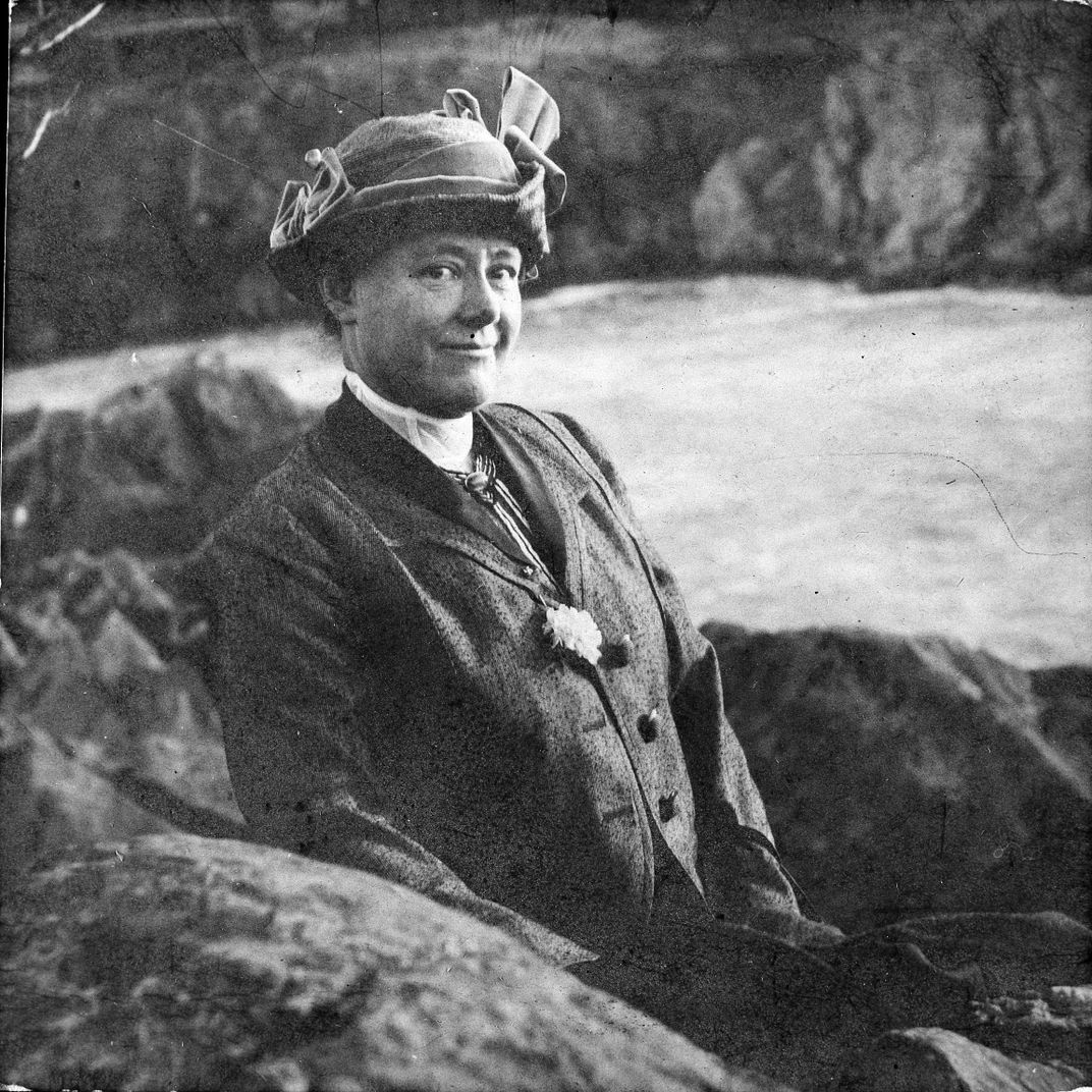 Black and white photo of Mary Vaux Walcott sitting on some rocks facing the camera, with waterfalls behind her