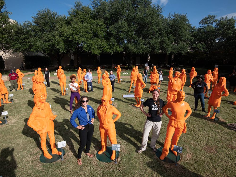 A field with life size orange plastic statues of women and two humans posing with them