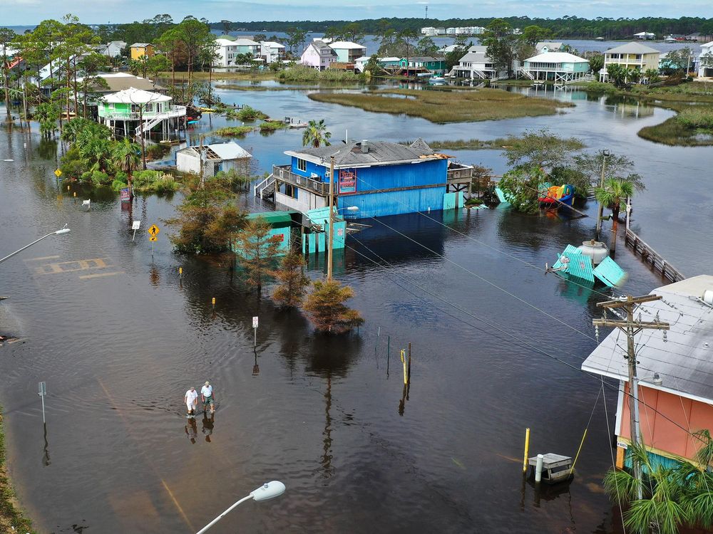 A flooded neighborhood from a bird's eye perspective, with two small figures walking in water that comes up to their calves. The street is entirely submerged, as are the foundations of the colorful houses