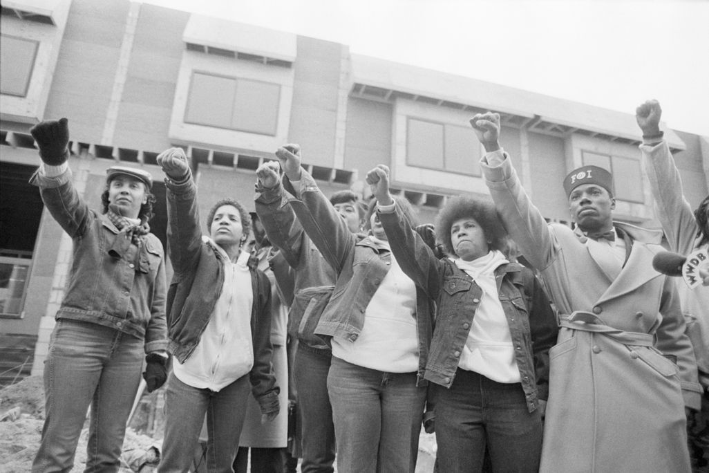 A black and white image of seven people, all Black, raising their right fists in the air in a line