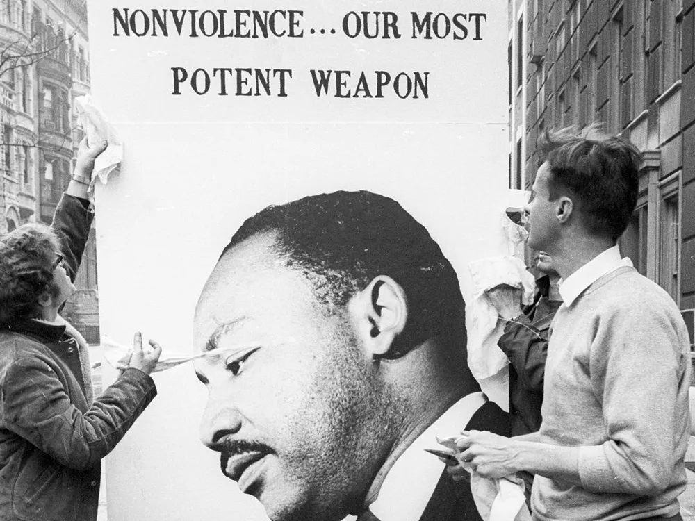 Two civilians look at a poster featuring the face of Martin Luther King Jr., his head depicted beneath the words 'Nonviolence... Our Most Potent Weapon.'