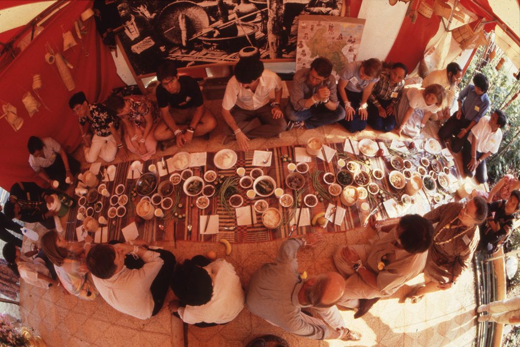 From above with a fisheye lens, a group of people gather around a long rectangular mat set with dishes of food, sitting on the ground.