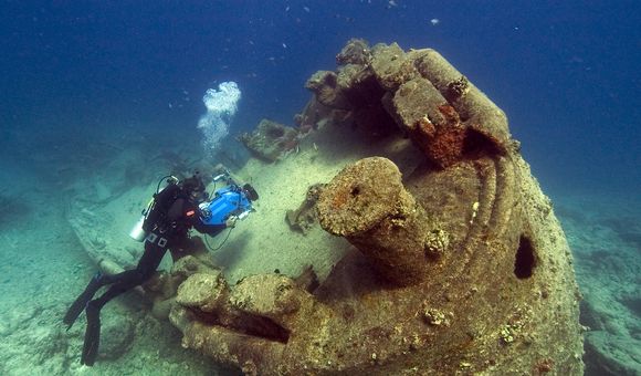 A diver uses a camera to study a barnacle-covered submarine resting on the seafloor