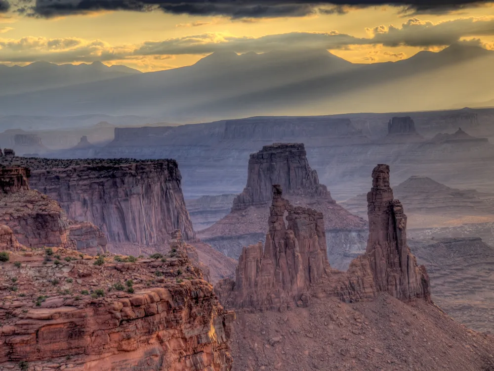 A landscape photo of Canyonlands National Park. The canyon is deep and dusty with tall, tower-like rocks jutting out of it. The sky is gold and gray, with wispy clouds.