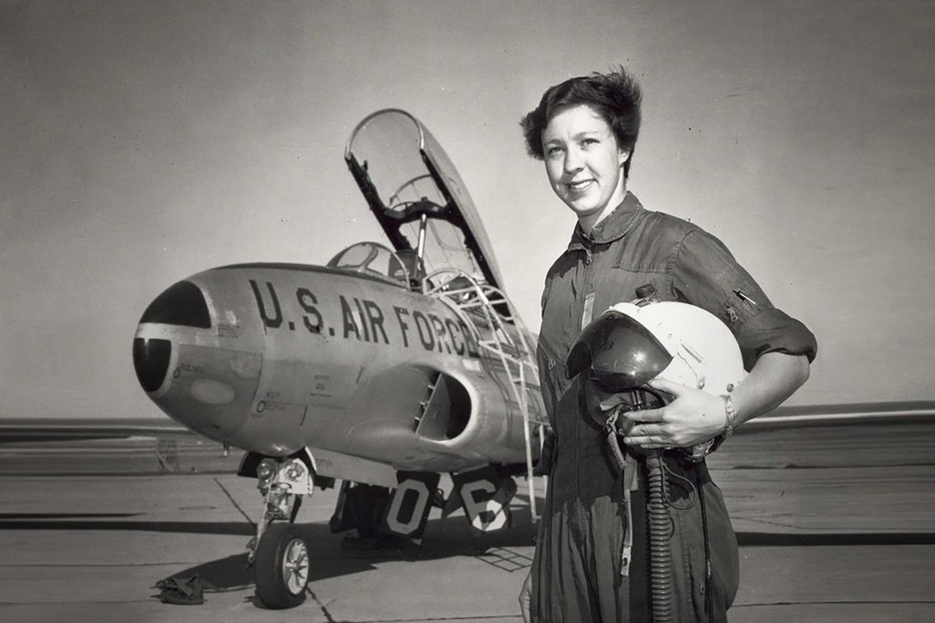 A photograph of a woman dressed to fly a jet, holding a helmet. The jet behind her has the top open and is labeled "U.S. Air Force"