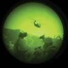 A grainy, green-tinted, circular view through a night vision scope shows a helicopter flying over a dark landscape at night.