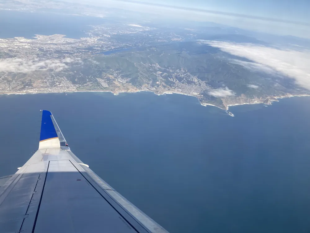 View from a plane’s window showing the wing as the plane flies over the ocean and shoreline.