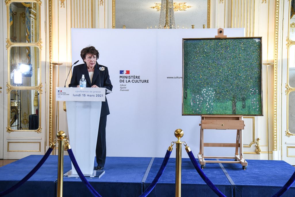 woman with brown hair and blue pantsuit standing at podium in front of pastel floral painting