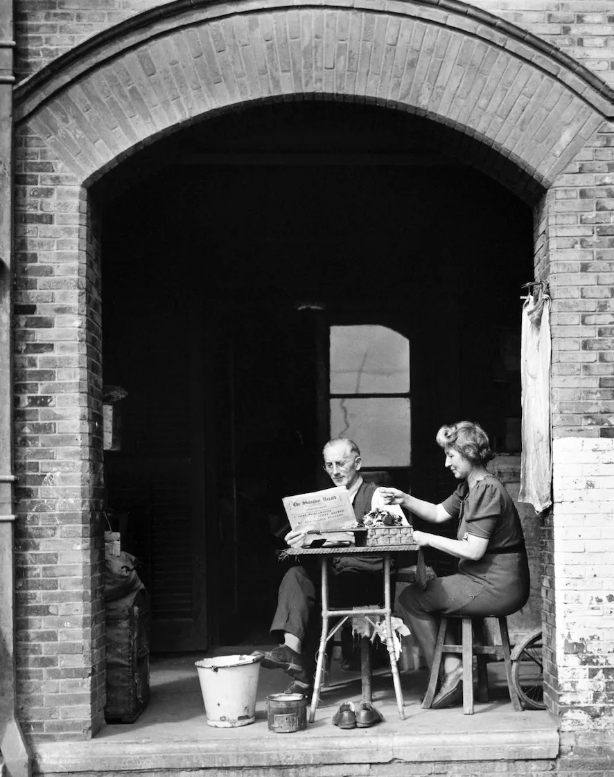 A man and woman sit at a small table in an archway; the man reads the paper and the woman pours a cup of tea