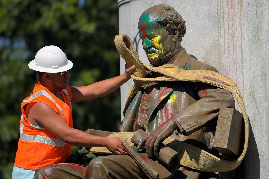 A man in a bright orange construction vest and white hat wraps a thick rope around a paint-splattered statue, of a bearded man wearing a suit and sitting in a chair