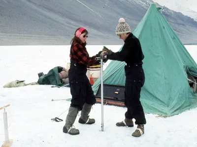 Eileen McSaveney (left) and Terry Tickhill (right) use a hand augur to drill Lake Vanda, Wright Valley, Antarctica, during the 1969-1970 field season. Water collected during this effort was used to date the lake.